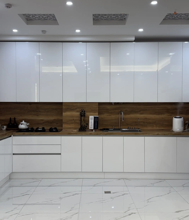 نمونه اجرای کابینت هایگلاس سفید براق با ترکیب مات و براق، صفحه و بین‌کابینتی طرح چوب، طراحی مدرن آشپزخانه توسط کابینت صحرایی با نورپردازی سقفی و چیدمان کلاسیک میز ناهارخوری